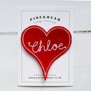 Puede incluir: Un parche bordado en forma de corazón rojo con el nombre "Chloe" en escritura cursiva blanca. El parche está delineado en blanco y se encuentra sobre una tarjeta blanca con borde verde. La tarjeta dice "PINE & BEAR Vintage Chainstitch".