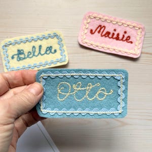 May include: Three rectangular felt name tags. One is yellow with "Bella" embroidered in blue, another is pink with "Maisie" in red, and the third is teal with "Otto" in cream. Each tag has a scalloped border.