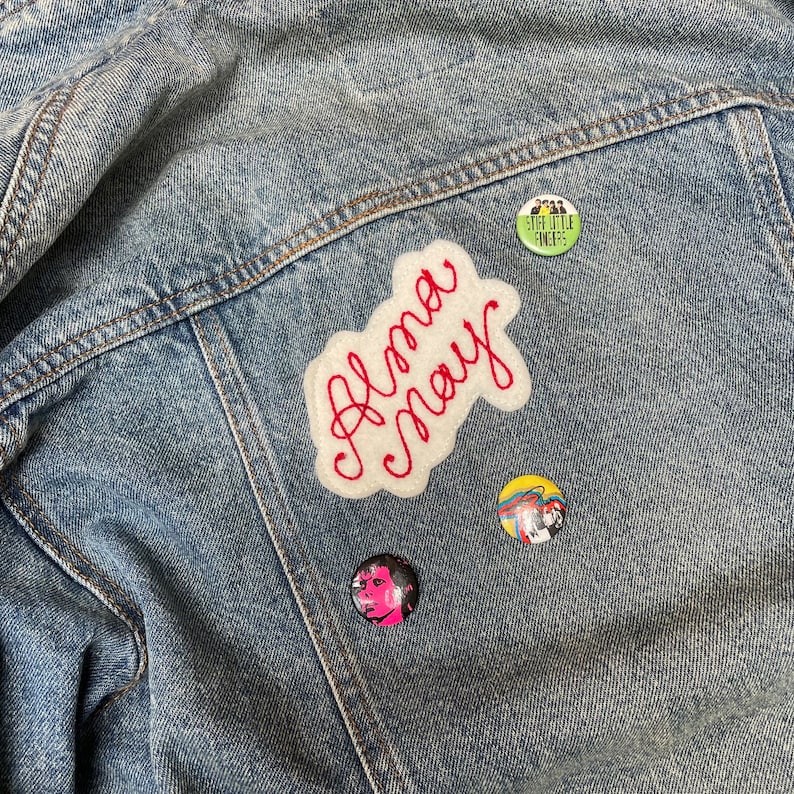 A denim jacket decorated with badges and a custom vintage chain stitch name patch in red and white.
