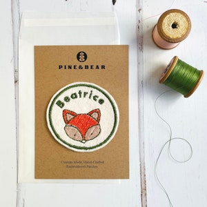 May include: A brown card with the text "PINE & BEAR" and "Custom-Made, Hand-Crafted Embroidered Patches."  A white circle with green stitching around the edge features a red fox with the name "Beatrice" embroidered in green.
