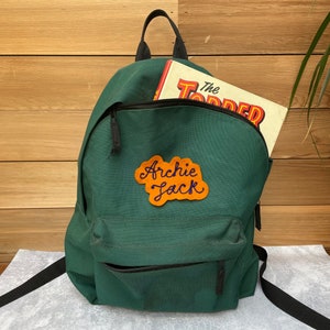 A green rucksack decorated with a custom vintage chain stitch name patch.