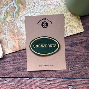 Puede incluir: Un parche bordado verde y dorado con la palabra "SNOWDONIA" en una tarjeta marrón con el logo "PINEANDBEAR" y la frase "Embroidered Patch". Un mapa y un objeto verde se encuentran en el fondo. El parche es ovalado.