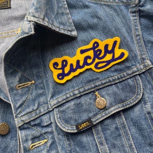 Puede incluir: Un parche de fieltro amarillo con la palabra "Lucky" bordada en hilo morado. El parche está cosido en una chaqueta vaquera azul.