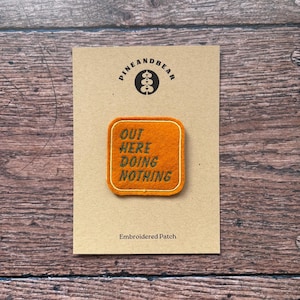 Puede incluir: Un parche bordado naranja con el texto "OUT HERE DOING NOTHING" en verde y amarillo. El parche es cuadrado con esquinas redondeadas y está sobre una tarjeta beige con el logotipo "PINEANDBEAR".