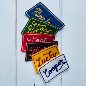 Puede incluir: Un conjunto de siete parches de fieltro con texto bordado. Los parches son de varios colores, incluyendo azul, verde, rojo, negro, amarillo y blanco. El texto en los parches incluye "Bonjour", "Stay Lucky", "Star", "Make", "Lucky", y "Cooper".
