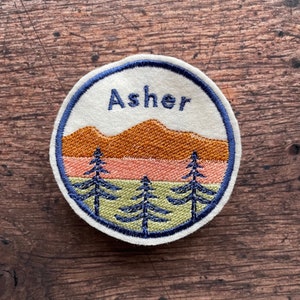 Puede incluir: Un parche redondo blanco bordado con un contorno azul. El parche presenta una cordillera en tonos naranja y rosa, con tres pinos verdes en frente. La palabra "Asher" está bordada en azul sobre la cordillera.