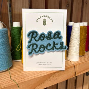 Pode incluir: Um patch de feltro azul claro com as palavras "Rosa Rocks" em escrita verde escuro. O patch está em um cartão branco com o logotipo "Pine and Bear" e o texto "Custom Chain Stitch Embroidered Patch". Bobinas de linha no fundo.