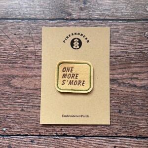 May include: Embroidered patch with the text "ONE MORE S'MORE" in a rectangular shape with a green border. The patch is on a tan card with the "PINEANDBEAR" logo. The words "Embroidered Patch" are printed below the patch.