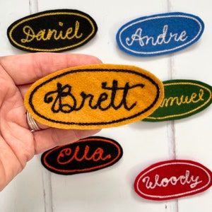 Puede incluir: Seis etiquetas de nombre ovaladas de fieltro con nombres bordados en hilo negro. Los nombres son Daniel, André, Brett, Samuel, Ella y Woody. Las etiquetas son de varios colores: negro, azul, amarillo, verde, naranja y rojo.