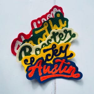May include: A collection of felt name patches in red, green, cream, yellow, and blue. Each patch displays a name in a contrasting colour, such as "Rocky" and "Austin". The patches are arranged on a white surface.