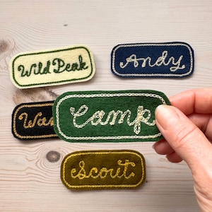 May include: A collection of embroidered felt patches in various colors, including green, navy, black, and olive. Each patch features white cursive text, with words like "Wild Peak," "Andy," "Camp," "War," and "Scout." The patches are rectangular with rounded corners.