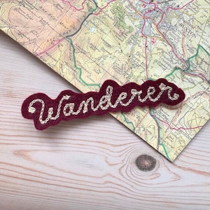 May include: A burgundy felt patch with the word "Wanderer" embroidered in white cursive script. The patch is placed on a vintage map, resting on a light wood surface. The map features place names and contour lines.