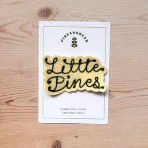 Pode incluir: Um patch de feltro amarelo com as palavras "Little Pines" em letras cursivas verde escuro. O patch está em um cartão branco com o logotipo "Pine and Bear" e o texto "Custom Chain Stitch Embroidered Patch".