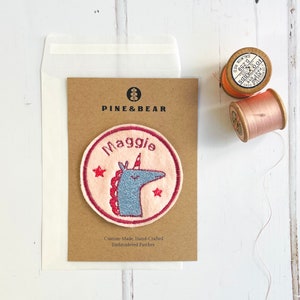 May include: A custom-made, hand-crafted embroidered patch featuring a unicorn with the name "Maggie". The patch is round with a pink background, a blue unicorn, and red stars. The text "PINE & BEAR" is at the top.