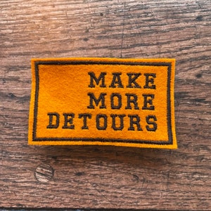 May include: A yellow felt patch with black embroidered text that reads "MAKE MORE DETOURS".