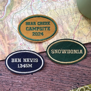 Puede incluir: Tres parches ovalados bordados. Uno es dorado con ribete verde y dice "NEAR CREEK CAMPSITE 2024". Otro es azul marino con ribete blanco y dice "BEN NEVIS 1345M". El tercero es verde con ribete dorado y dice "SNOWDONIA".