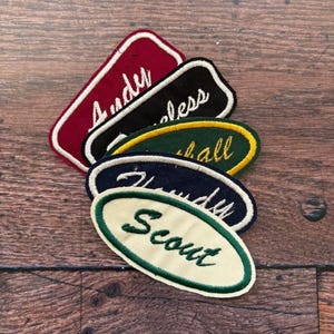 May include: A selection of embroidered name patches in different shapes and colours. The patches display names such as "Andy", "less", "Marshall", "Randy", and "Scout" in cursive lettering. The patches are in shades of red, black, green, and navy blue.