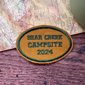May include: An oval patch with the text "BEAR CREEK CAMPSITE 2024" embroidered in green on a gold background, with a green border. The patch is placed on a map and a wooden surface.