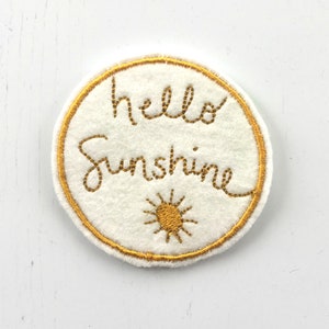 May include: A white felt circle with a gold embroidered border and the text "hello sunshine" in gold thread. A small gold embroidered sun is below the text.