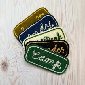 May include: A collection of embroidered felt patches in shades of green, navy, and black. Each patch features a white stitched border and embroidered text, including "Camp" and "Andy". The patches are rectangular with rounded corners.