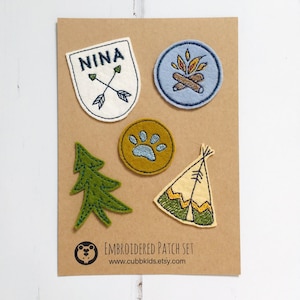 May include: Embroidered patch set with a shield reading "NINA" and crossed arrows, a campfire, a paw print, a pine tree, and a teepee. The patches are in white, blue, green, and yellow, on a brown card. The set is for decorative purposes.