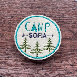 May include: Circular embroidered patch in off-white, featuring the words "CAMP SOFIA" in teal and dark blue. It has a teal border, an arrow, and three green embroidered pine trees. A great accessory for camping.