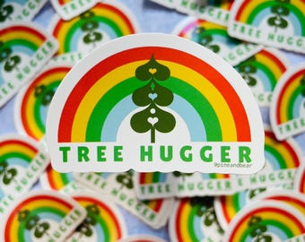 May include: A sticker with a rainbow arch and a green tree with hearts in the center. The text "TREE HUGGER" is written in green letters below the rainbow. The text "@pineandbear" is written in smaller letters below the text "TREE HUGGER".
