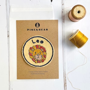May include: A brown card with the text "PINE&BEAR" and "Leo" in a circle with a lion embroidered on it. The card says "Custom-Made, Hand-Crafted Embroidered Patches".
