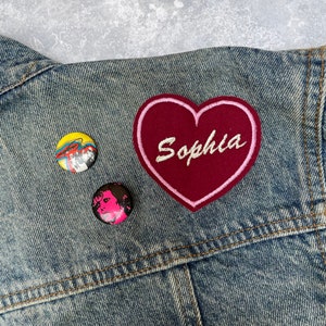 Könnte beinhalten: Eine Jeansjacke mit einem rosa Herz-Patch, auf dem "Sophia" in weißer Stickerei steht. Auf der Jacke befinden sich zwei kleine runde Knöpfe mit bunten Designs.