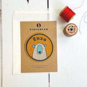 May include: A brown card with the text "PINE & BEAR" and "Enzo" in a yellow circle with a white embroidered bear inside. The card is on a white wooden surface with a spool of red thread and a spool of blue thread.