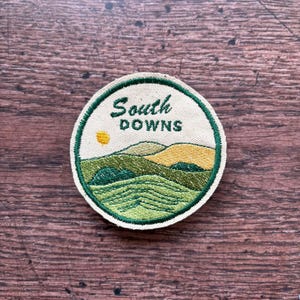 May include: Circular embroidered patch with the words "South Downs" in green. The design shows a landscape of green hills, yellow accents, and a yellow sun. It has a green border and a beige background, ideal for jackets or bags.