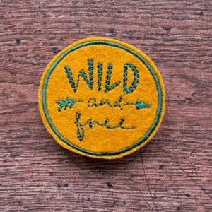 May include: A round, mustard-yellow patch with the words "WILD and free" embroidered in green. The text is inside a green circle, with an arrow design. The patch is made of a felt-like material.