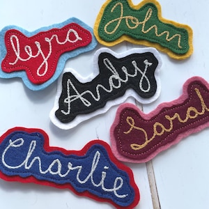 Puede incluir: Cinco etiquetas de nombre de fieltro con nombres bordados en diferentes colores. Los nombres son "Lyra", "John", "Andy", "Sarah" y "Charlie".