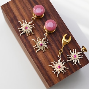 May include: Two pairs of pink and gold ear gauges with a sun and moon design. The gauges are made of glass and metal.