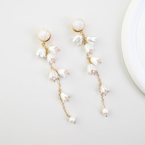 May include: A pair of delicate earrings with a gold chain, white flower and pearl details. The earrings have a textured white ball at the top. The earrings dangle and are set against a white background.