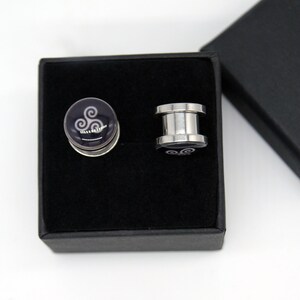 May include: A pair of silver ear gauges with a black and white swirl design in a black velvet box.