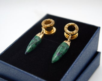 Puede incluir: Un par de dilatadores de oreja de color dorado con detalles de piedra verde. Los dilatadores se muestran en una caja de terciopelo azul.