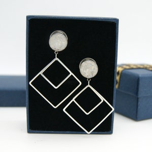 May include: A pair of silver earrings with a white stone and a geometric design. The earrings are displayed in a black box with a blue lid.
