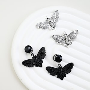 May include: An assortment of butterfly-shaped jewelry. Two silver-colored butterfly pendants and two pairs of black butterfly earrings with black circular studs are displayed on a white, ridged surface. The butterflies have detailed wing patterns.