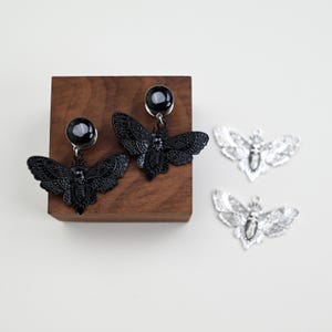 May include: Black moth-shaped earrings with a dark gem and silver moth pendants. The earrings are displayed on a wooden block. Two silver moth pendants are also visible, offering a contrasting metallic accent.