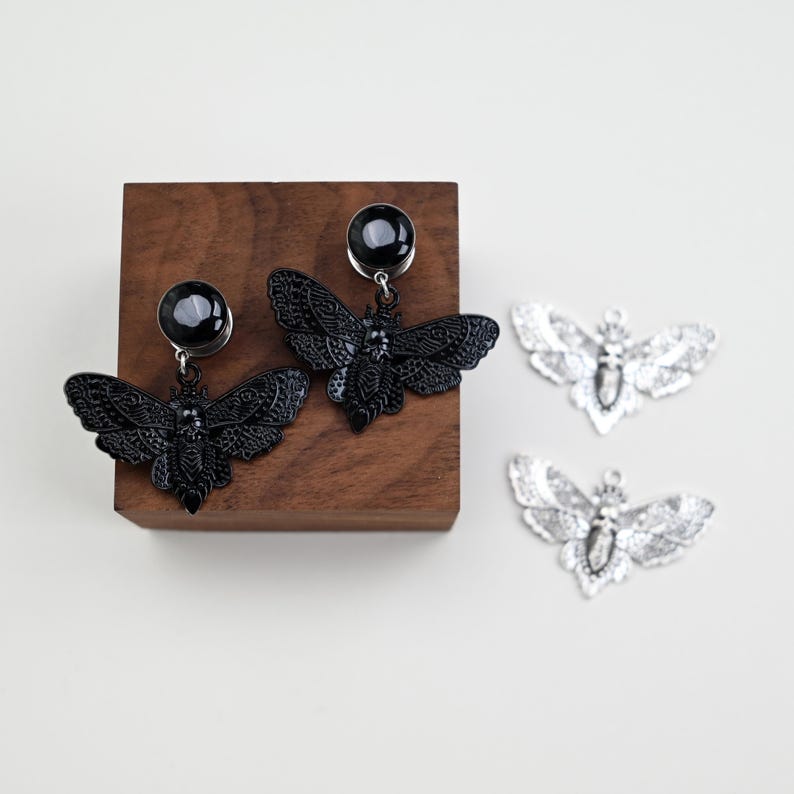 May include: Black moth-shaped earrings with a dark gem and silver moth pendants. The earrings are displayed on a wooden block. Two silver moth pendants are also visible, offering a contrasting metallic accent.