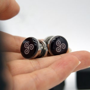 May include: A pair of silver magnetic earrings with a black background and a white triquetra symbol.