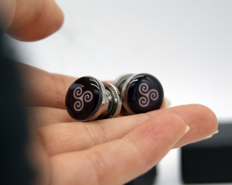 May include: A pair of silver magnetic earrings with a black background and a white triquetra symbol.