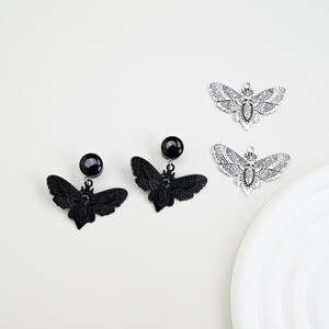 May include: Black and silver-toned moth-shaped earrings and charms. Two pairs of black earrings with a round stud and a moth pendant are displayed alongside two silver-toned moth charms. The moth wings are detailed with intricate patterns.