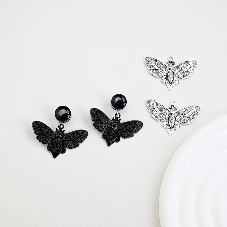 May include: Black and silver-toned moth-shaped earrings and charms. Two pairs of black earrings with a round stud and a moth pendant are displayed alongside two silver-toned moth charms. The moth wings are detailed with intricate patterns.