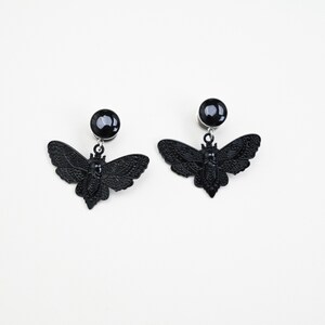 May include: A pair of black earrings featuring a moth design. Each earring has a round, black stud and a detailed moth pendant. The moth pendants have intricate wing patterns and are suspended from the studs.