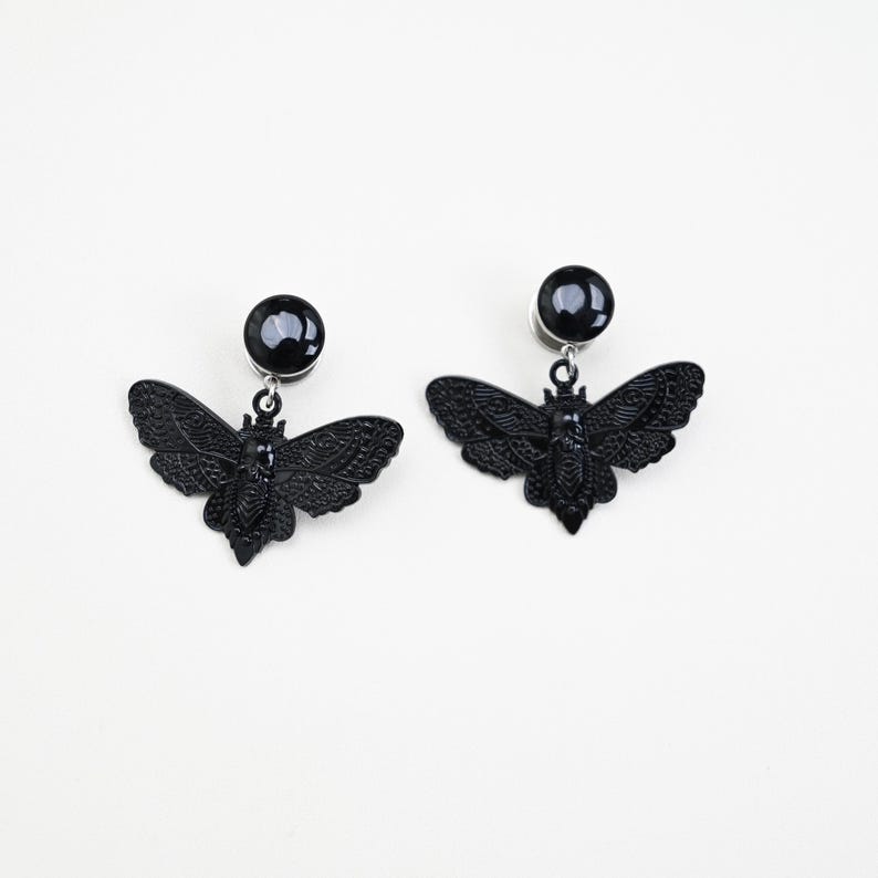 May include: A pair of black earrings featuring a moth design. Each earring has a round, black stud and a detailed moth pendant. The moth pendants have intricate wing patterns and are suspended from the studs.