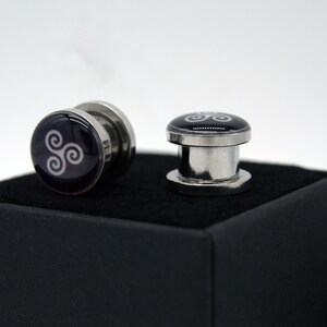 May include: Two silver ear gauges with a black background and a white Celtic knot design.