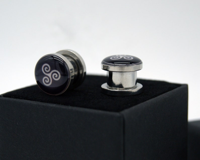 May include: Two silver ear gauges with a black background and a white Celtic knot design.