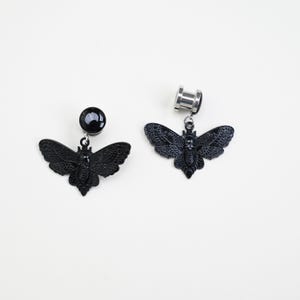 May include: Two black butterfly-shaped ear gauges. One gauge has a black, round end, while the other has a silver, cylindrical end. Both have a detailed butterfly pendant hanging from them. The butterflies have intricate wing patterns.